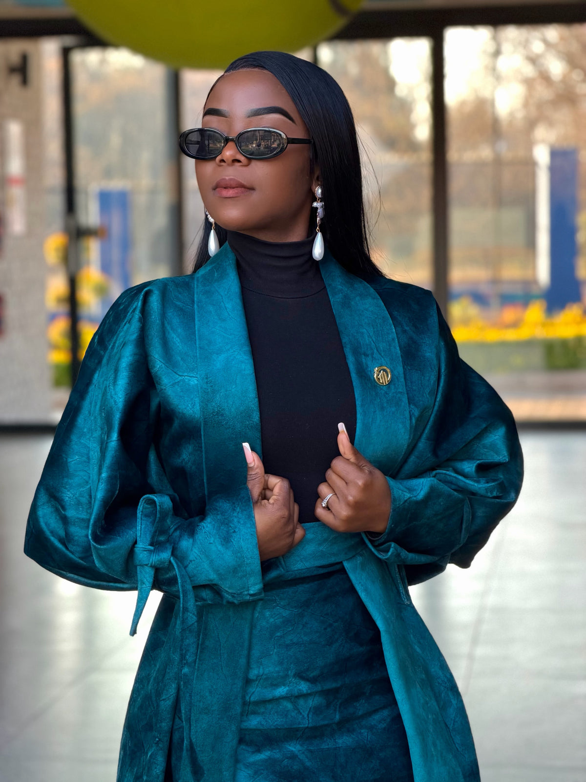 Close-up of a teal textured belted coat with tie-cuff sleeves, worn over a black turtleneck