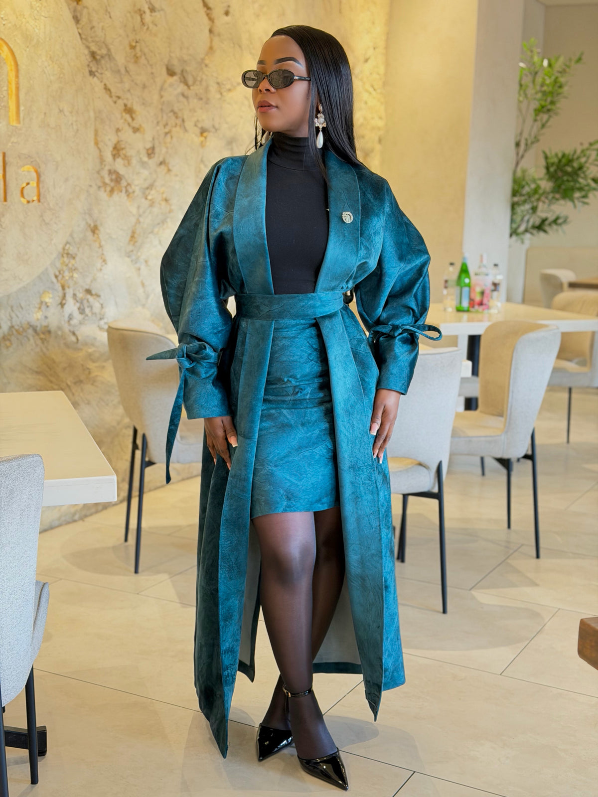 A woman wearing a teal blue satin-like long coat over a black turtleneck, styled with black pointed shoes and sunglasses, standing in a modern cafe interior.